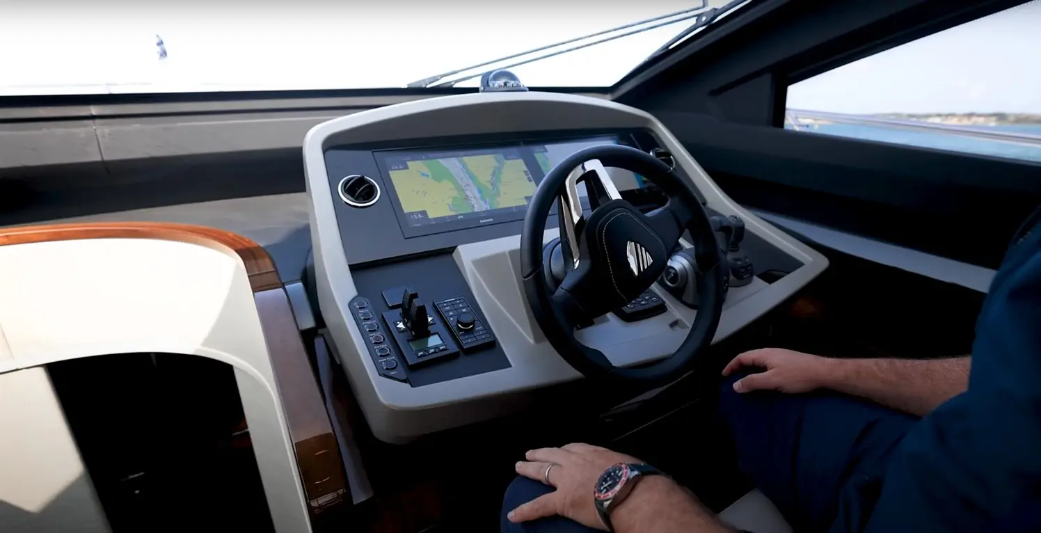 Modern yacht cockpit showing a steering wheel, navigation screen, and controls, with a hand resting on the armrest.